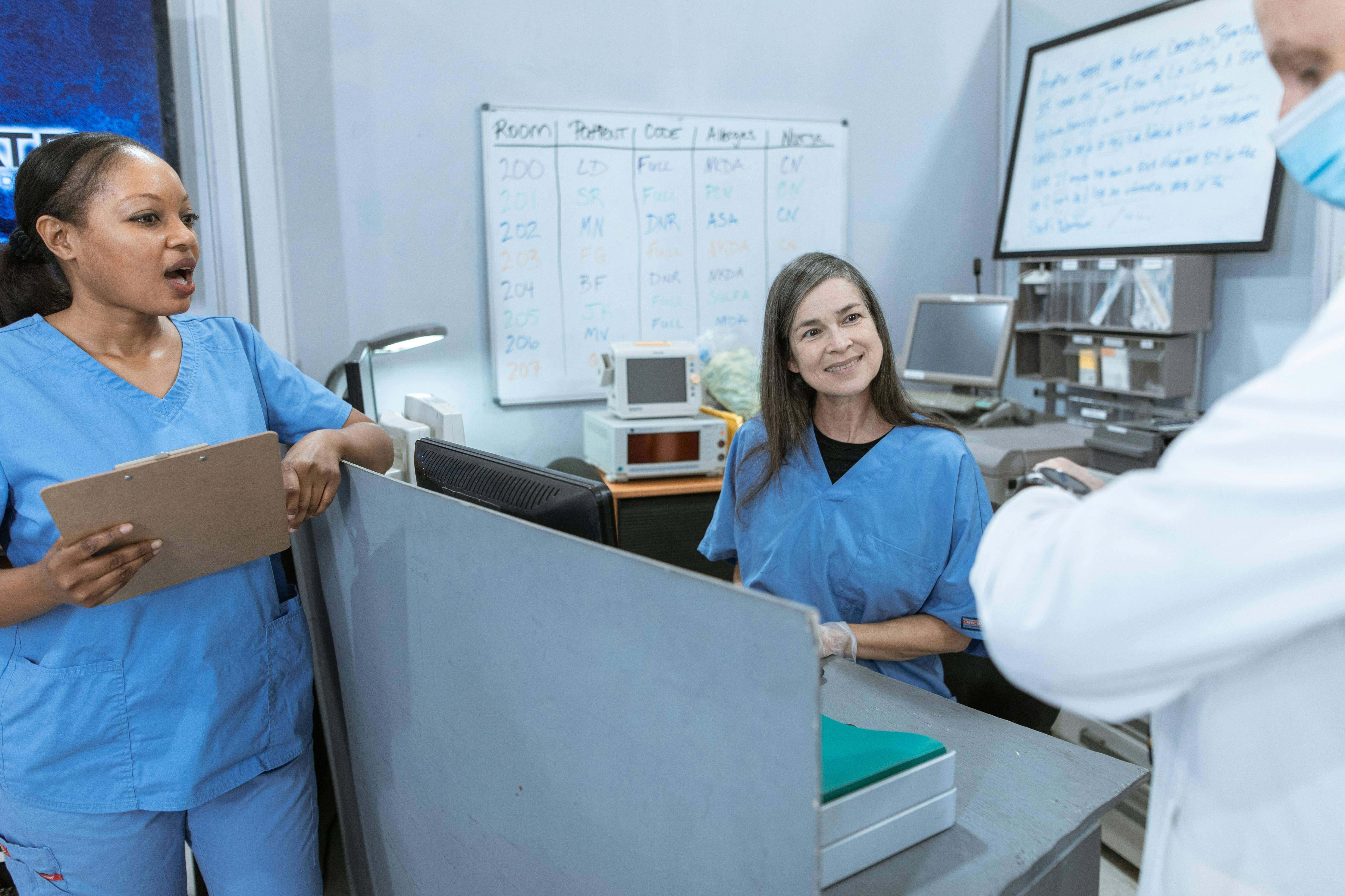 A doctor checks watch while speaking to a nurse with clipboard and another nurse at a nurse's station